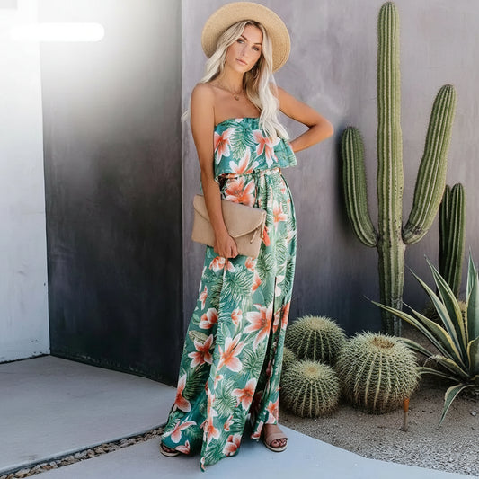 A woman wearing a tropical print strapless dress with a maxi length, paired with a straw hat and carrying a beige purse.
