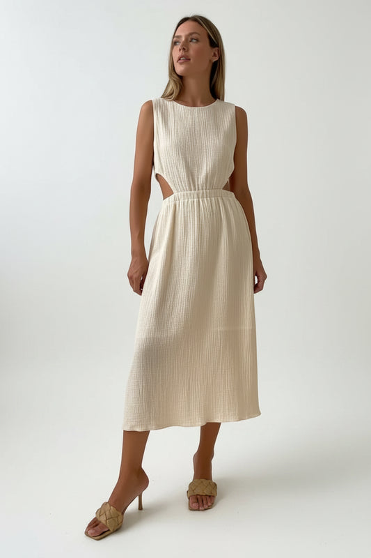 A woman standing against a white background, wearing a beige, sleeveless, mid-calf long dress with a V-neckline and button details.