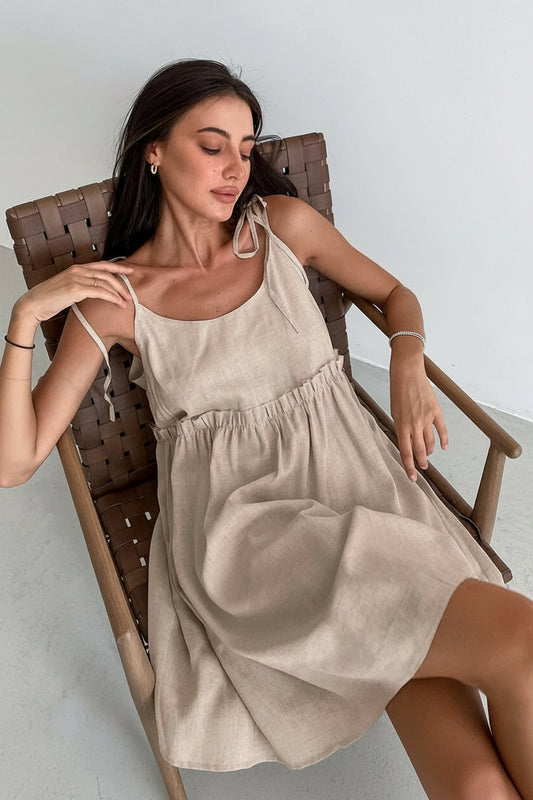 A woman sitting in a wooden chair wearing a beige spaghetti strap A-line mini dress with ruched details.