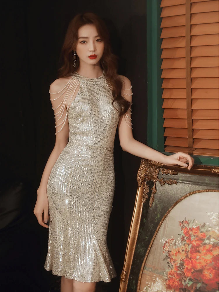 A woman wearing a sparkling silver sequined dress with shoulder chain details, posing next to an ornate gold picture frame in front of wooden shutters.