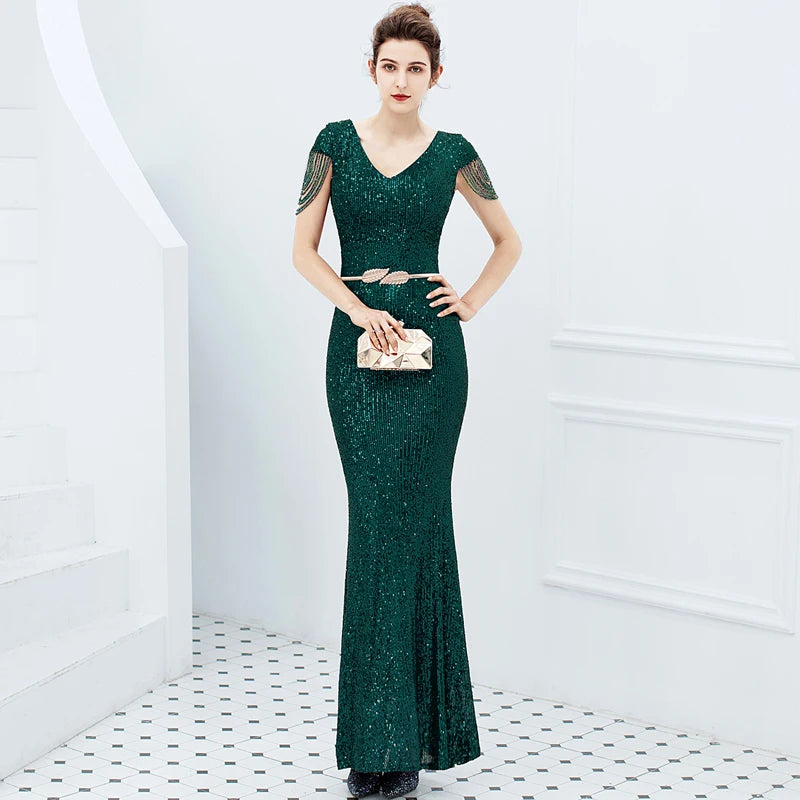 A woman models a sparkling dark green sequined mermaid gown with sheer short sleeves and a matching belt, holding a metallic clutch in a bright white interior setting.