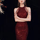 A woman wearing a sparkling deep red sequined halter-neck gown with shoulder strap details, posed against a black background.