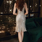 A woman stands with her back to the camera wearing a shimmering champagne sequin midi dress with sheer long sleeves and a flared hem, posed in front of a window with string lights and a dark green velvet sofa.
