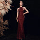 A woman models a floor-length burgundy sequined gown with a high neckline and asymmetrical shoulder cutouts, posed against a dark backdrop with dried floral decor on the left.