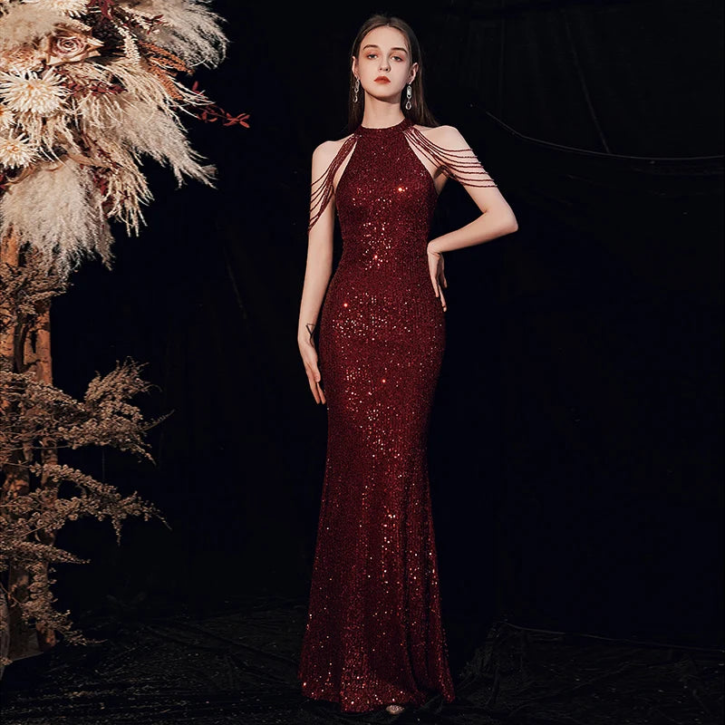 A woman models a floor-length burgundy sequined gown with a high neckline and asymmetrical shoulder cutouts, posed against a dark backdrop with dried floral decor on the left.