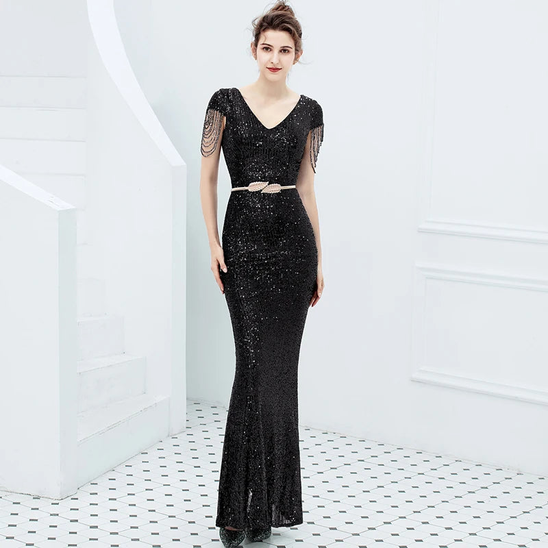 A woman models a black sequined evening gown with sheer flutter sleeves, a V-neckline, and a thin metallic belt at the waist, set against a bright white interior background with a tiled floor.