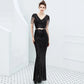 A woman models a black sequined evening gown with sheer flutter sleeves, a V-neckline, and a thin metallic belt at the waist, set against a bright white interior background with a tiled floor.