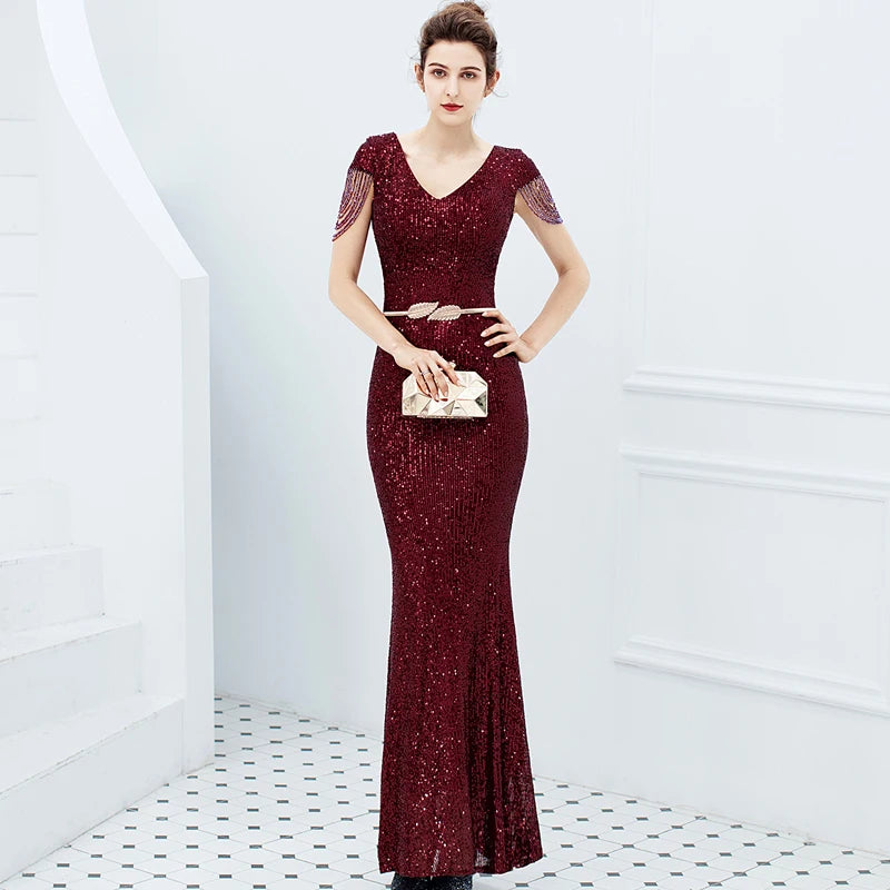 A woman models a floor-length burgundy sequined evening gown with short sheer sleeves, a V-neckline, and a matching belt, holding a small metallic clutch in a bright white interior setting.