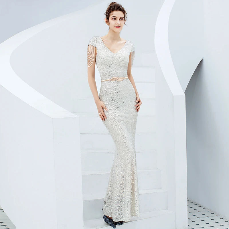 A woman stands on white architectural stairs wearing a fitted champagne-ivory evening gown with textured beaded sleeves and a matching belt detail.