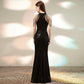 A woman stands with her back to the camera wearing a floor-length black sequined evening gown with cold-shoulder straps and a mermaid silhouette, photographed against a softly lit neutral wall.