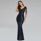 A woman modeling a floor-length navy blue sequined gown with a V-neckline, sheer cap sleeves with fringe detailing, and a fitted mermaid silhouette, set against a solid light gray studio background.