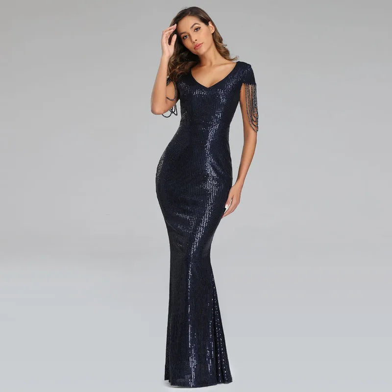 A woman modeling a floor-length navy blue sequined gown with a V-neckline, sheer cap sleeves with fringe detailing, and a fitted mermaid silhouette, set against a solid light gray studio background.