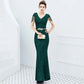 A woman models a sparkling dark green sequined mermaid gown with sheer short sleeves and a matching belt, holding a metallic clutch in a bright white interior setting.