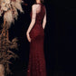 A woman wearing a deep burgundy sequined evening gown with a backless design and double shoulder straps, posed against a dark background with dried floral decorations.
