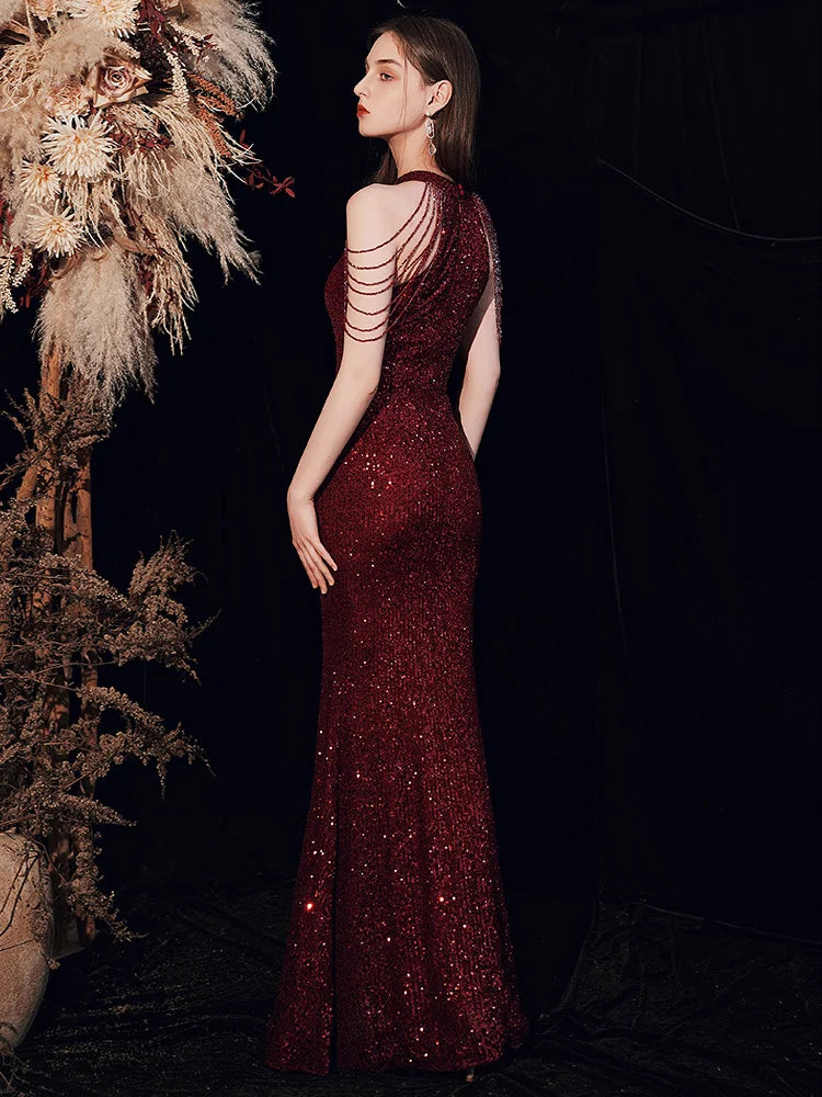 A woman wearing a deep burgundy sequined evening gown with a backless design and double shoulder straps, posed against a dark background with dried floral decorations.