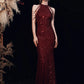 A woman stands against a black backdrop wearing a floor-length, sleeveless burgundy sequined gown with halter-style straps and fringe details. Pampas grass decor is visible on the left side of the frame.