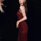 A woman modeling a floor-length, deep burgundy sequined gown with a single strapless neckline and multiple thin metallic side straps, posed against a dark background with dried floral decor at the upper left.
