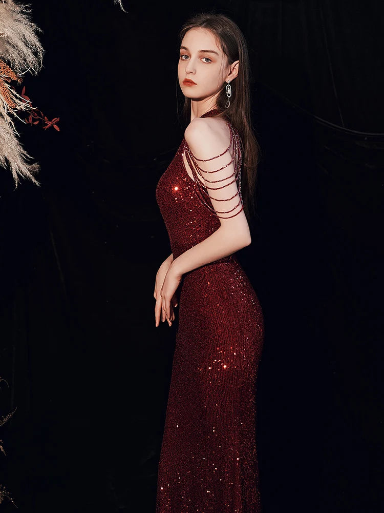 A woman modeling a floor-length, deep burgundy sequined gown with a single strapless neckline and multiple thin metallic side straps, posed against a dark background with dried floral decor at the upper left.