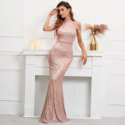 A woman models a floor-length, high-neck sequined gown in a pale blush pink tone. The dress features thin shoulder straps and a fitted silhouette that flares into a mermaid-style hem.