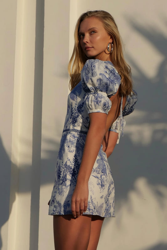 A woman modeling a short puff-sleeve dress with a blue and white toile pattern, photographed outdoors against a sunlit white wall with leaf shadows.