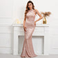 A woman models a rose gold sequined mermaid gown with sheer draped shoulder straps, standing in front of a white fireplace mantel decorated with candles and a gold vase.