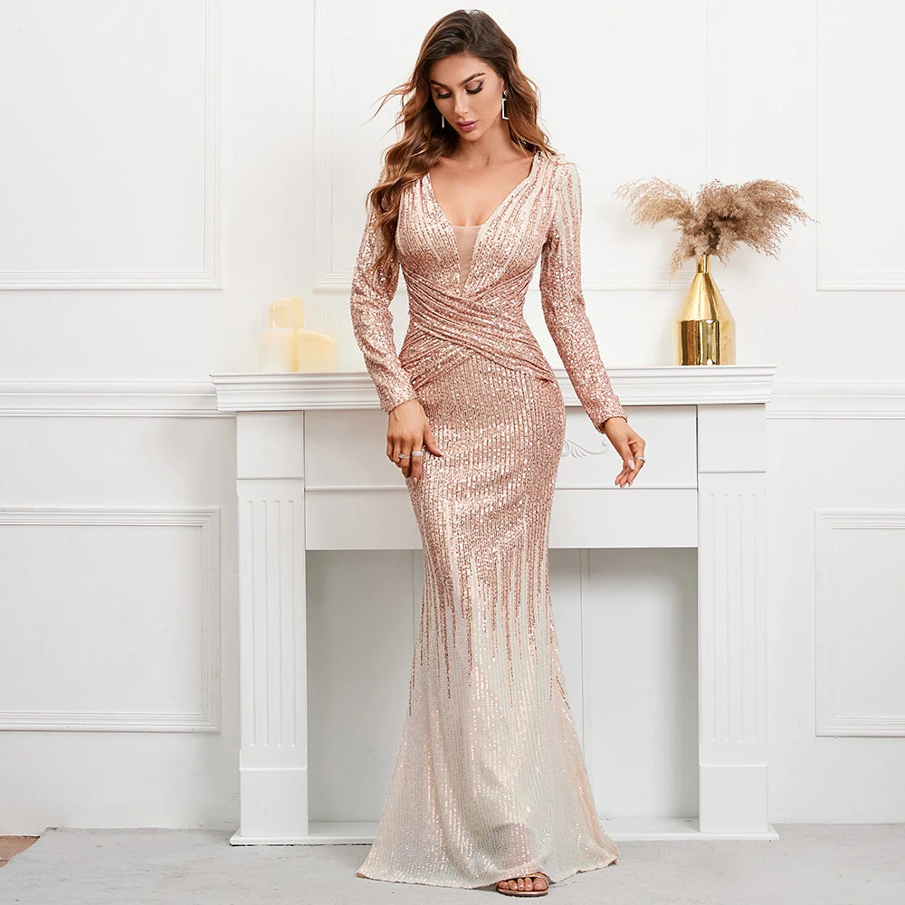 A woman models a long-sleeve, floor-length champagne pink sequined gown with a V-neckline, standing in front of a white fireplace mantel in a bright, modern room.