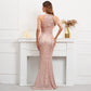 A woman stands with her back to the camera wearing a floor-length rose gold sequined gown with halter-style shoulder straps and a fitted mermaid silhouette, posed in front of a white fireplace mantel in an indoor room.