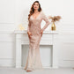A woman models a long-sleeve, deep V-neck floor-length gown covered in shimmering rose gold sequins, standing in front of a white fireplace mantel.