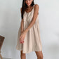 A woman sitting in a wooden chair wearing a beige spaghetti strap A-line mini dress with ruched details.
