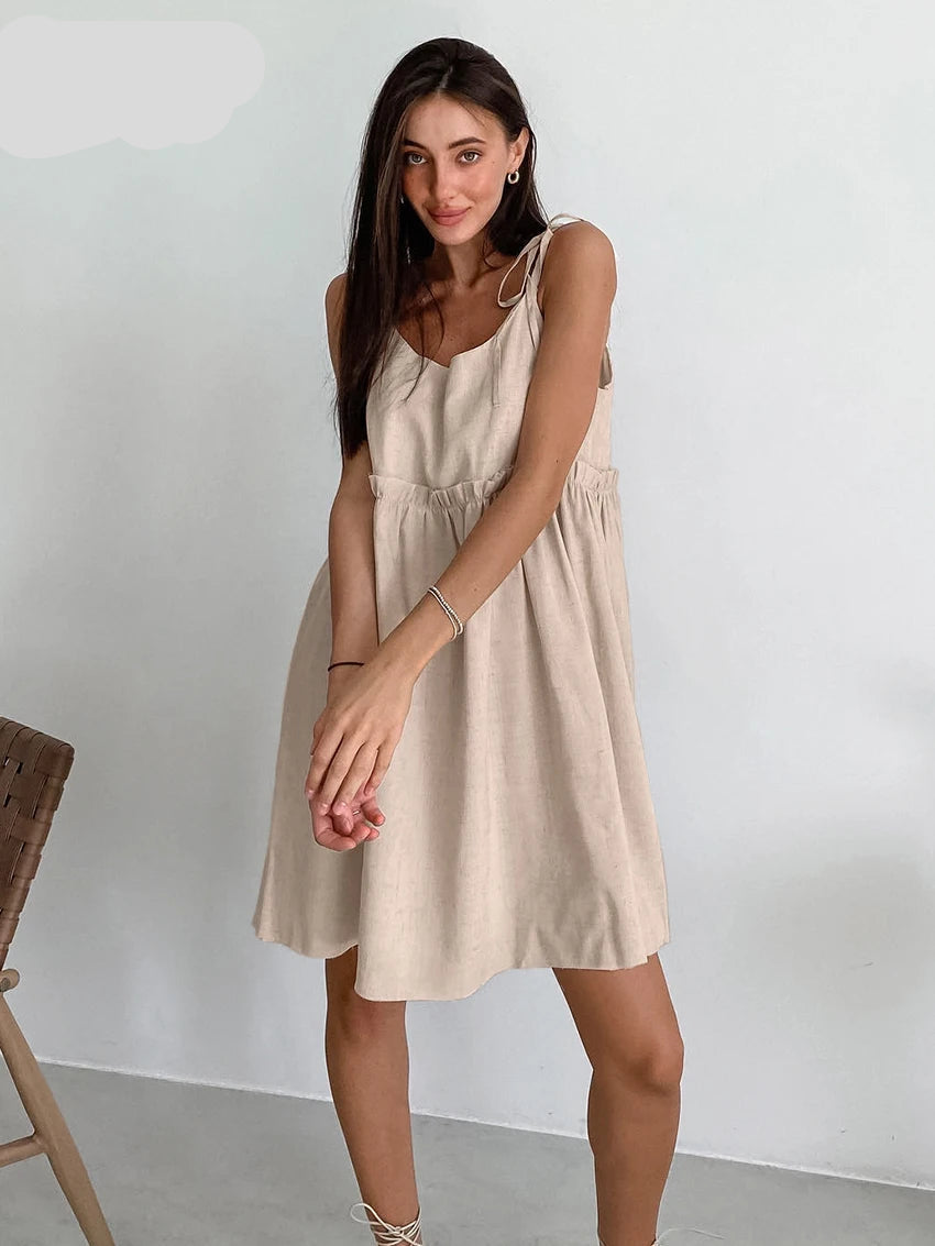 A woman sitting in a wooden chair wearing a beige spaghetti strap A-line mini dress with ruched details.