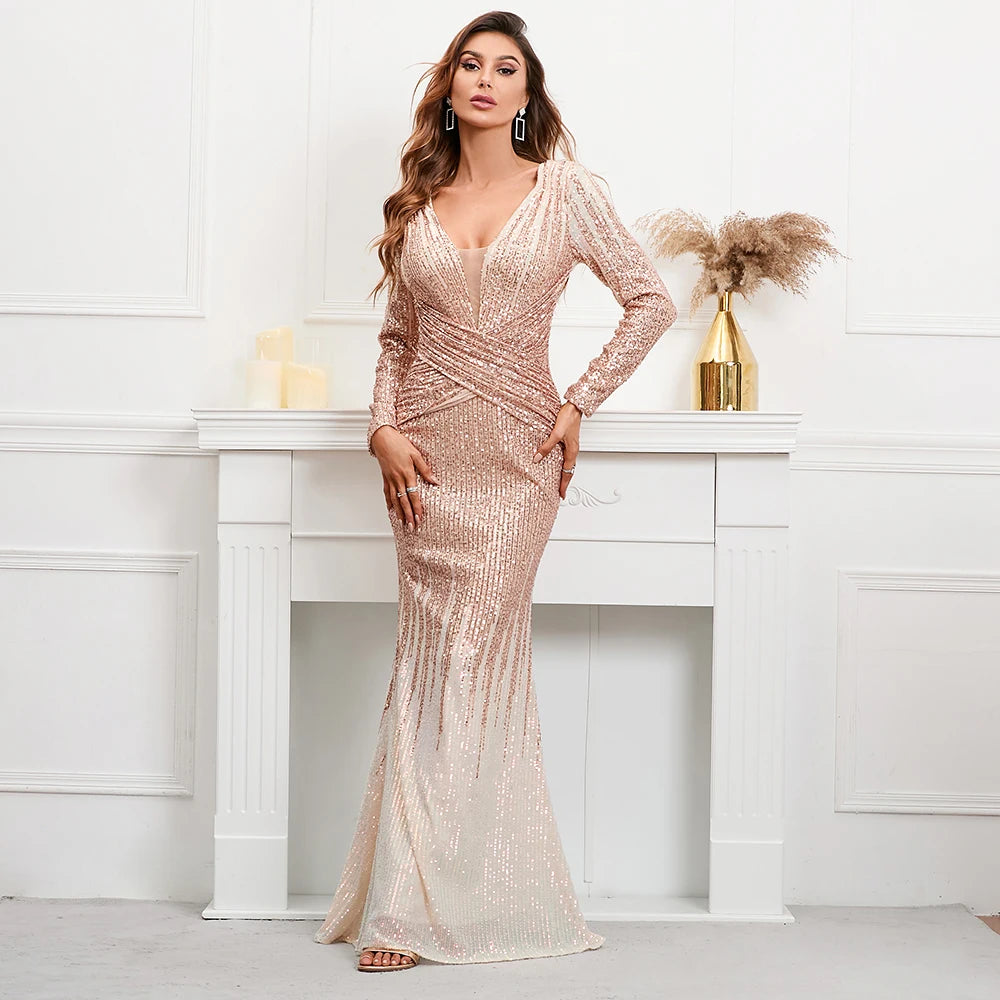 A woman models a long-sleeve, deep V-neck floor-length gown covered in shimmering rose gold sequins, standing in front of a white fireplace mantel in a bright room.