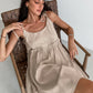 A woman sitting in a wooden chair wearing a beige spaghetti strap A-line mini dress with ruched details.