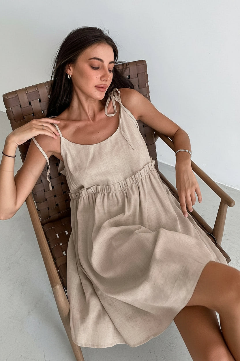 A woman sitting in a wooden chair wearing a beige spaghetti strap A-line mini dress with ruched details.