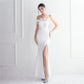 A woman models a strapless off-the-shoulder white evening gown with a fitted bodice, sheer illusion neckline, high thigh slit, and flared hem, paired with sparkly silver heels against a white paneled wall.