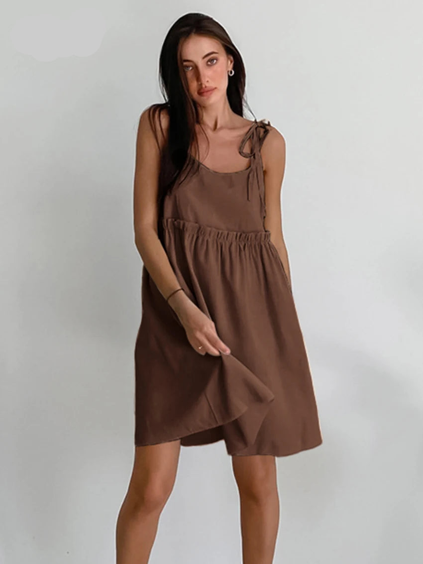 A woman sitting in a wooden chair wearing a beige spaghetti strap A-line mini dress with ruched details.