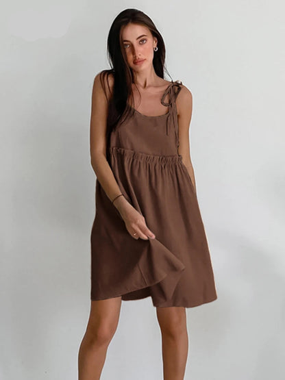 A woman sitting in a wooden chair wearing a beige spaghetti strap A-line mini dress with ruched details.